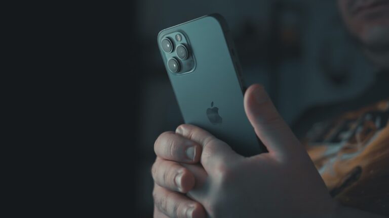 A close-up of a person holding a modern iPhone, with the camera lens array clearly visible, set against a dark, moody background.