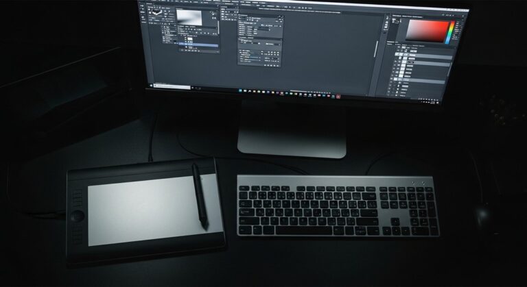 An overhead view of a professional designer's dark workspace, showing a monitor with Adobe Photoshop open, highlighting the complexity of a solo creative's workflow.